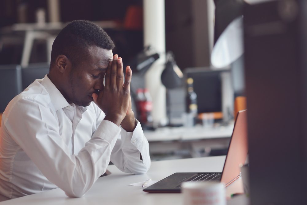 Tired African man sitting at a office after a hard workday, working on laptop, trying to concentrate