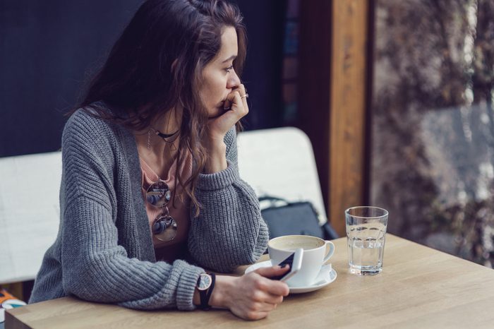 woman holding her phone, looking through the window and drinking coffee