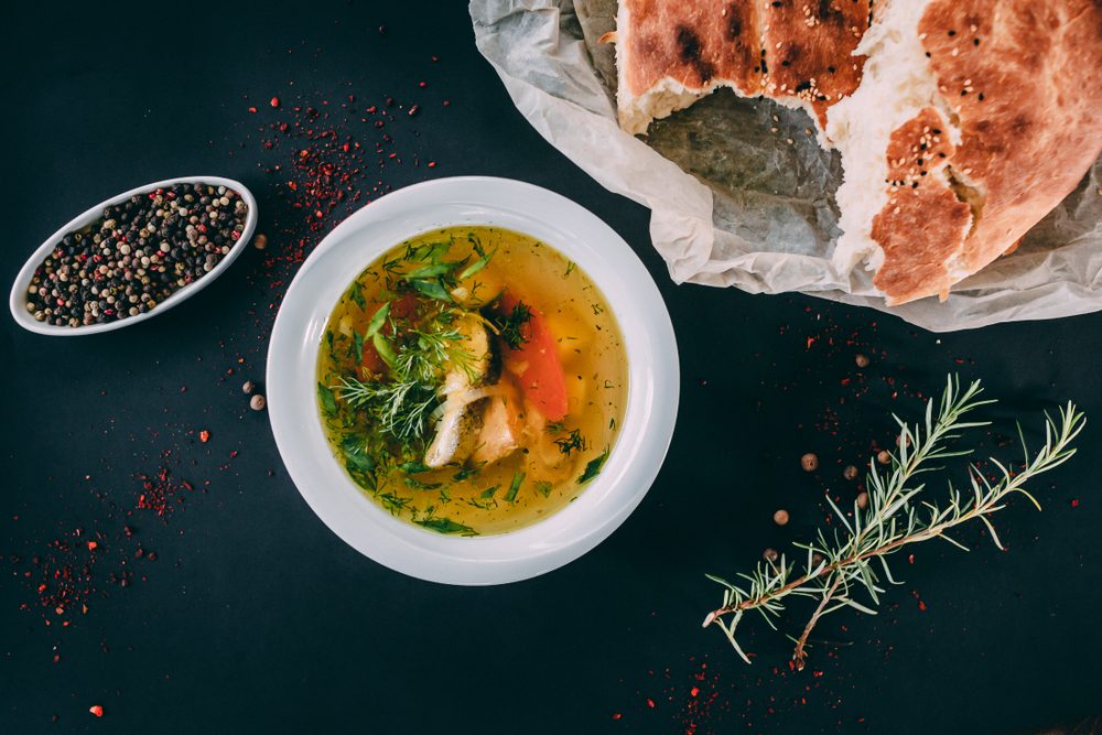 Ukha russian fish soup with trout, parsley, herbs and vegetables in it and a ground pepper nearby with rosemary, crushed pepper and a fresh bread on the craft paper on the dark background.