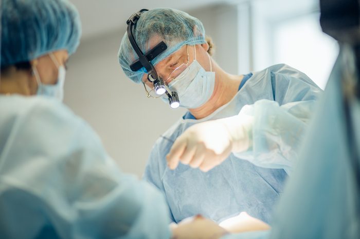 Surgeon performing cosmetic surgery on breasts in hospital operating room. Surgeon in mask wearing surgical loupes during medical procedure. Breast augmentation, enlargement, enhancement