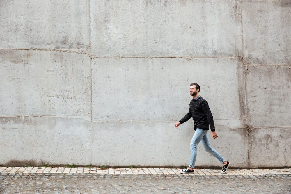 Full length portrait of a smiling casual man walking on a city street against gray wall