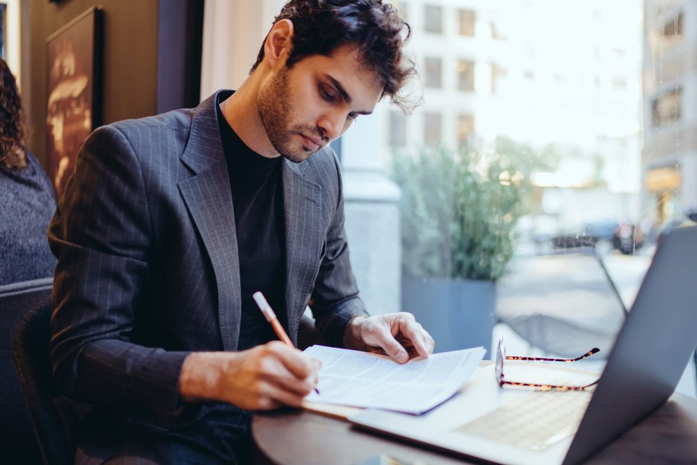 Young handsome caucasian businessman  in trendy suit writing information on financial papers for banking on laptop computer, serious confident male doing paperwork remotely indoors in coffee shop 