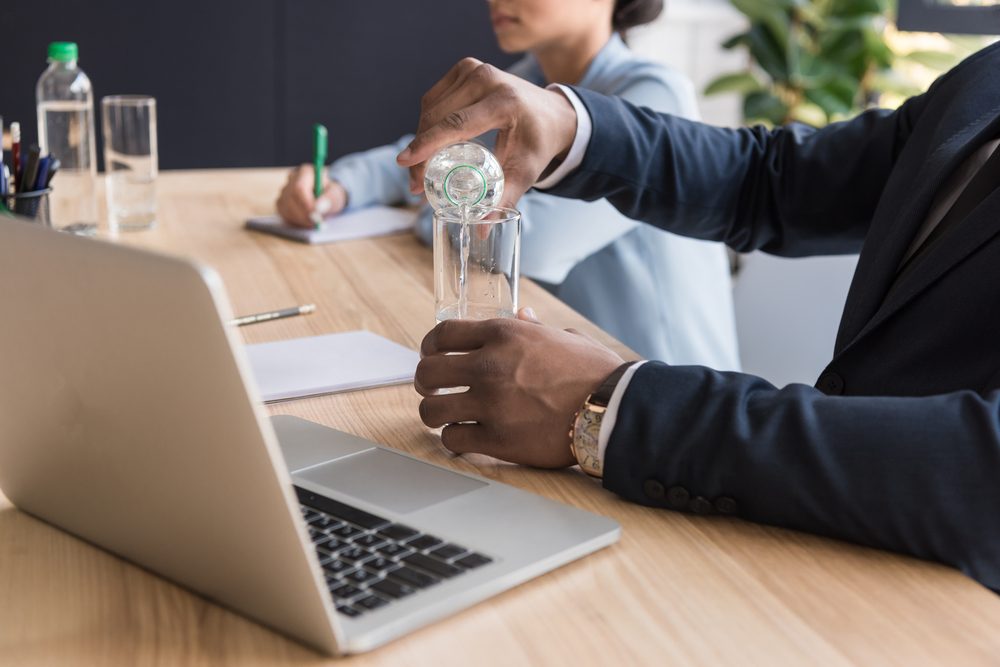 partial view of african american businessman pouring water into glass at workplace with laptop