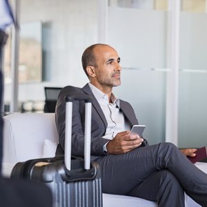 Mature businessman expecting airplane at the airport. Thoughtful business man waiting for flight in airport. Formal business man sitting in airport waiting room with luggage and phone in hand.