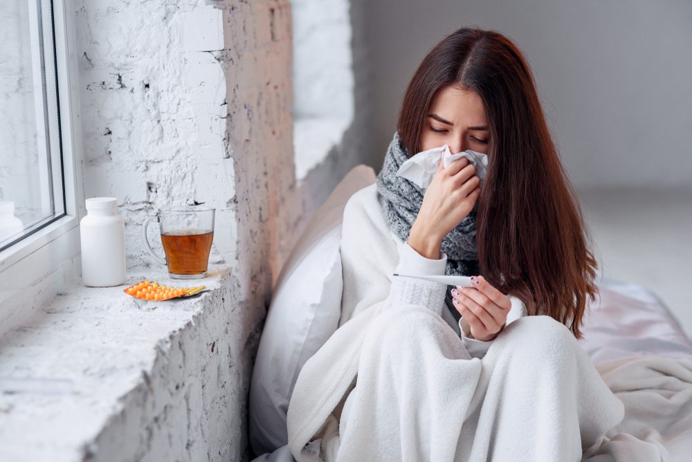 Cold and flu. Sick woman caught cold, feeling illness and sneezing in paper wipe. Closeup of beautiful unhealthy girl covered in blanket wiping nose and looking at thermometer. Healthcare concept.