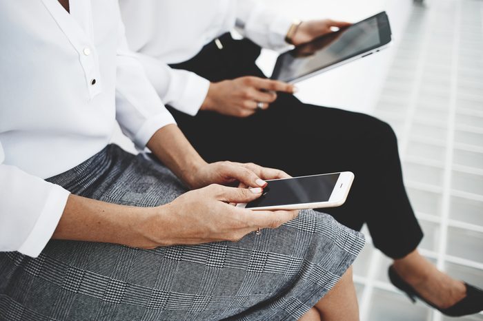 Closeup of woman is typing text message on mobile phone, while female near her is watching video on touch pad. Business women are booking on-line plane ticket via cell telephone and digital tablet 