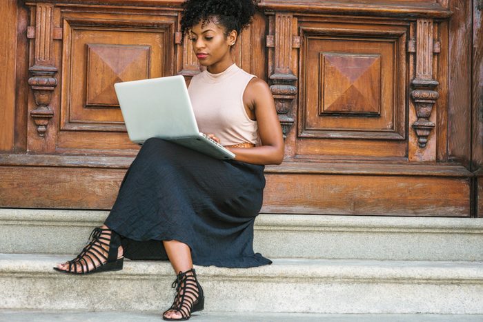 African American Student studying in New York, with afro hairstyle, wearing sleeveless light color top, black skit, strappy sandals, sitting by office door in New York, working on laptop computer.