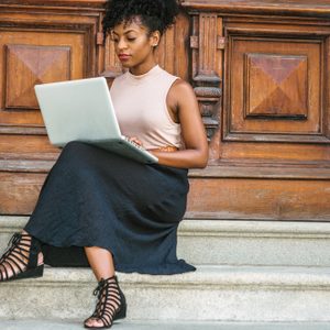 African American Student studying in New York, with afro hairstyle, wearing sleeveless light color top, black skit, strappy sandals, sitting by office door in New York, working on laptop computer.