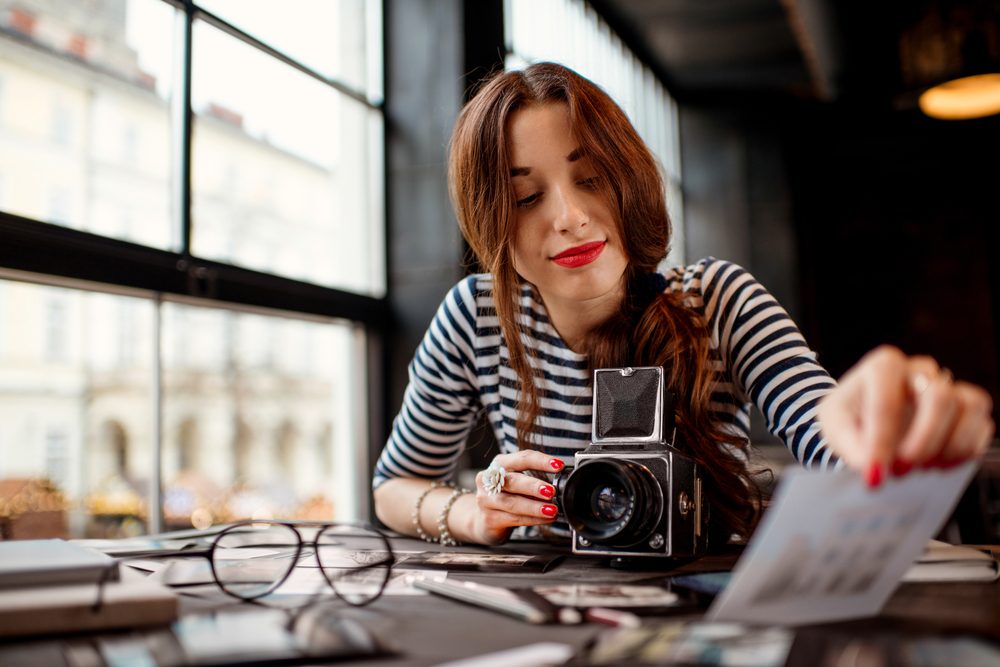 Young woman photographer looking at the printed photos with old 6x6 frame camera sitting in the cafe with loft design interior