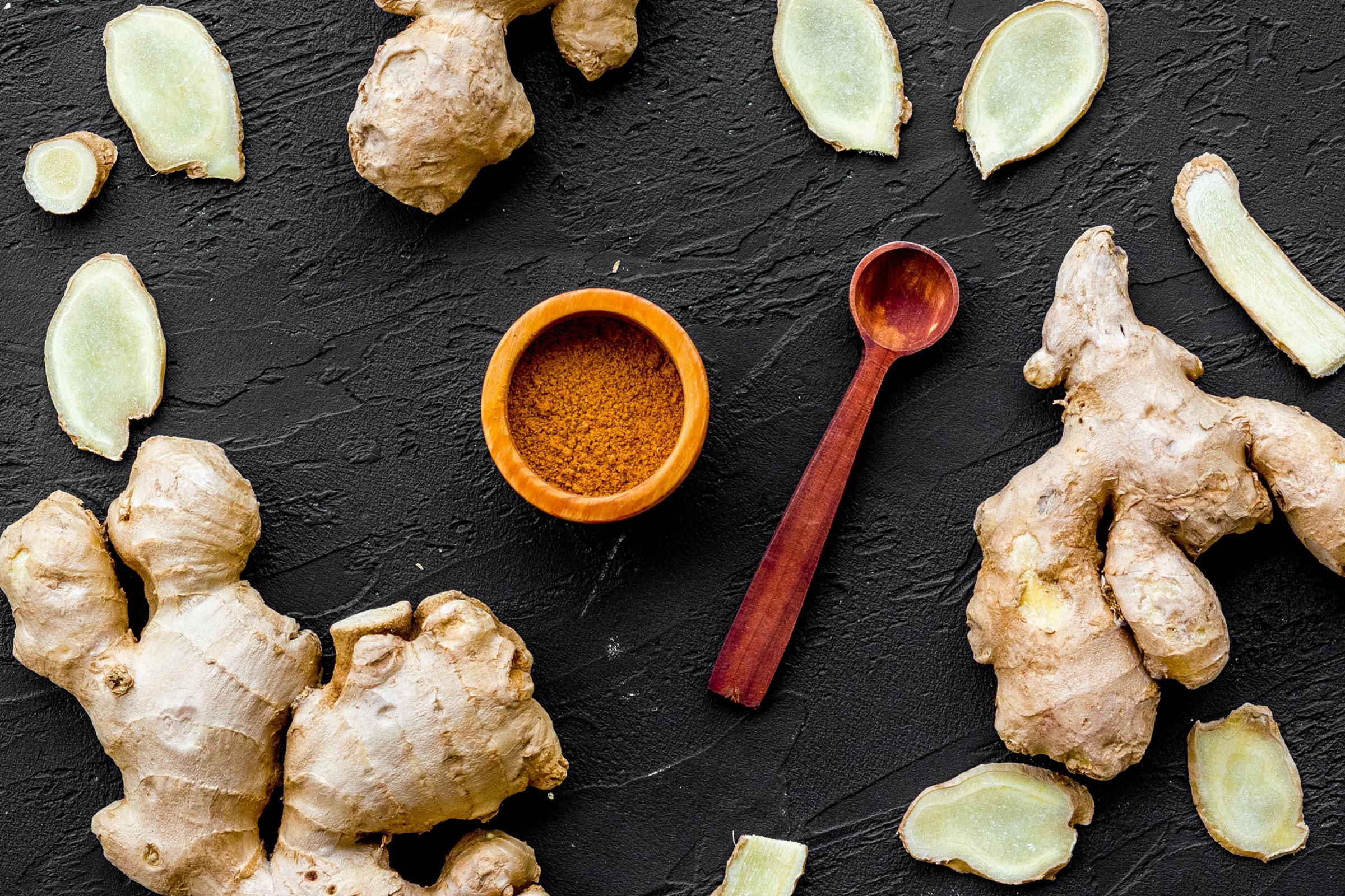 Seasoning. Ground ginger in small bowl near sliced ginger root on black background top view