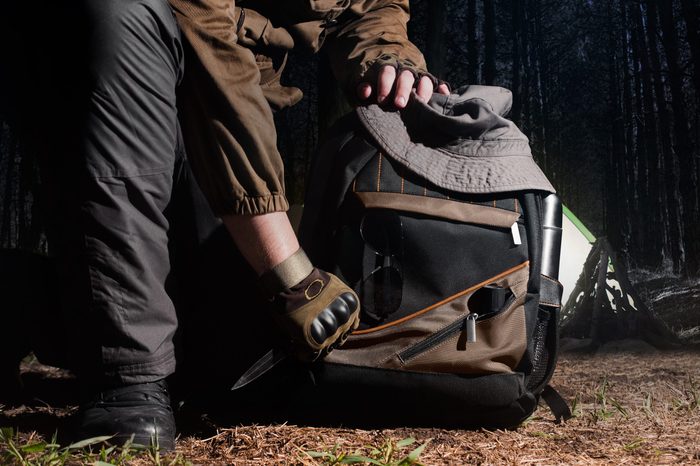 Man in tactical outfit holding a knife and kneeling for backpack with camping and tactical gear on night forest background.