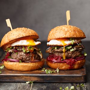 Two homemade beef burgers with mushrooms, micro greens, red onion, fried eggs and beet sauce on wooden cutting board