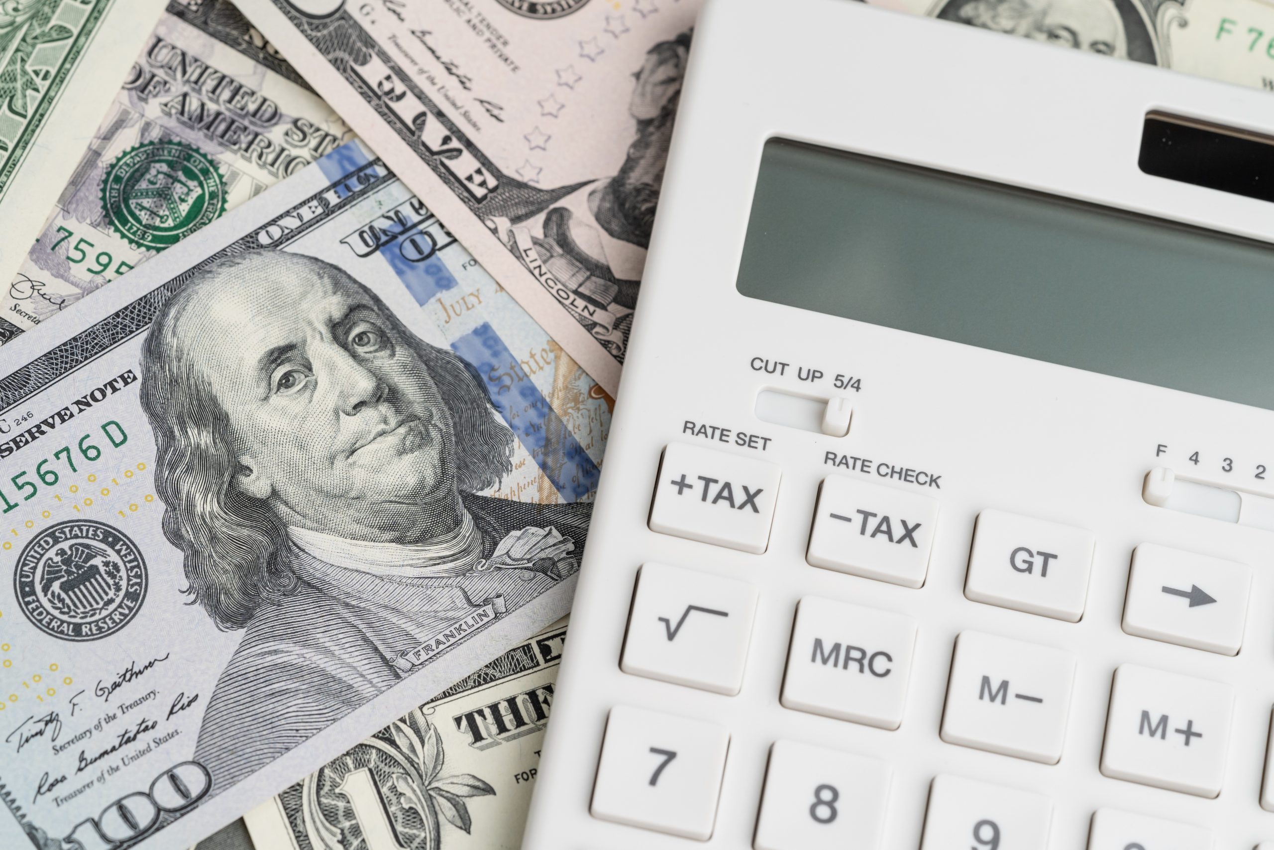 Flat lay or top view of clean white calculator with tax plus and tax minus buttons on pile of US Dollar banknotes money using as tax calculation, budget, investment or financial activities concept.