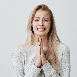 Beautiful blonde female clenching teeth with begging look holding palms pressed together in front of her, asking for forgiveness, feeling sorry after she hurt her boyfriend's feelings. Body language