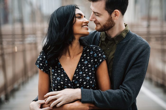 Love story in New York. Gorgeous couple of American man with beard and tender Eastern woman hug each other before the cityscape of Brooklyn bridge somewhere in New York