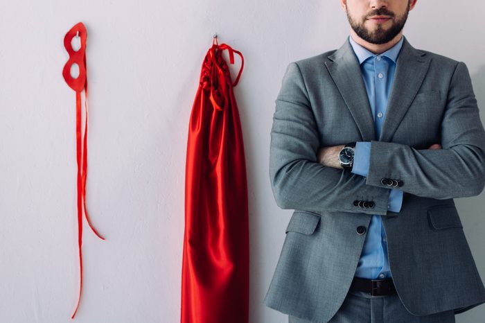 cropped image of super businessman standing with crossed arms near mask and cape on wall in office 