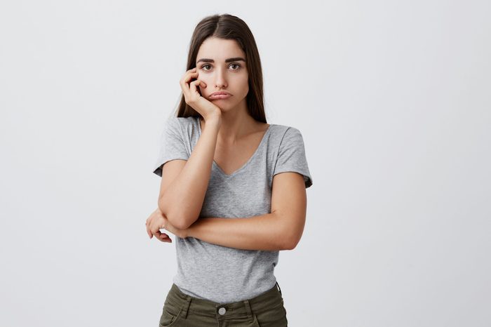 Portrait of young sad charming caucasian female student with dark long hair in stylish gray outfit holding head with hand, looking in camera with unhappy expression after receiving bad mark on exam