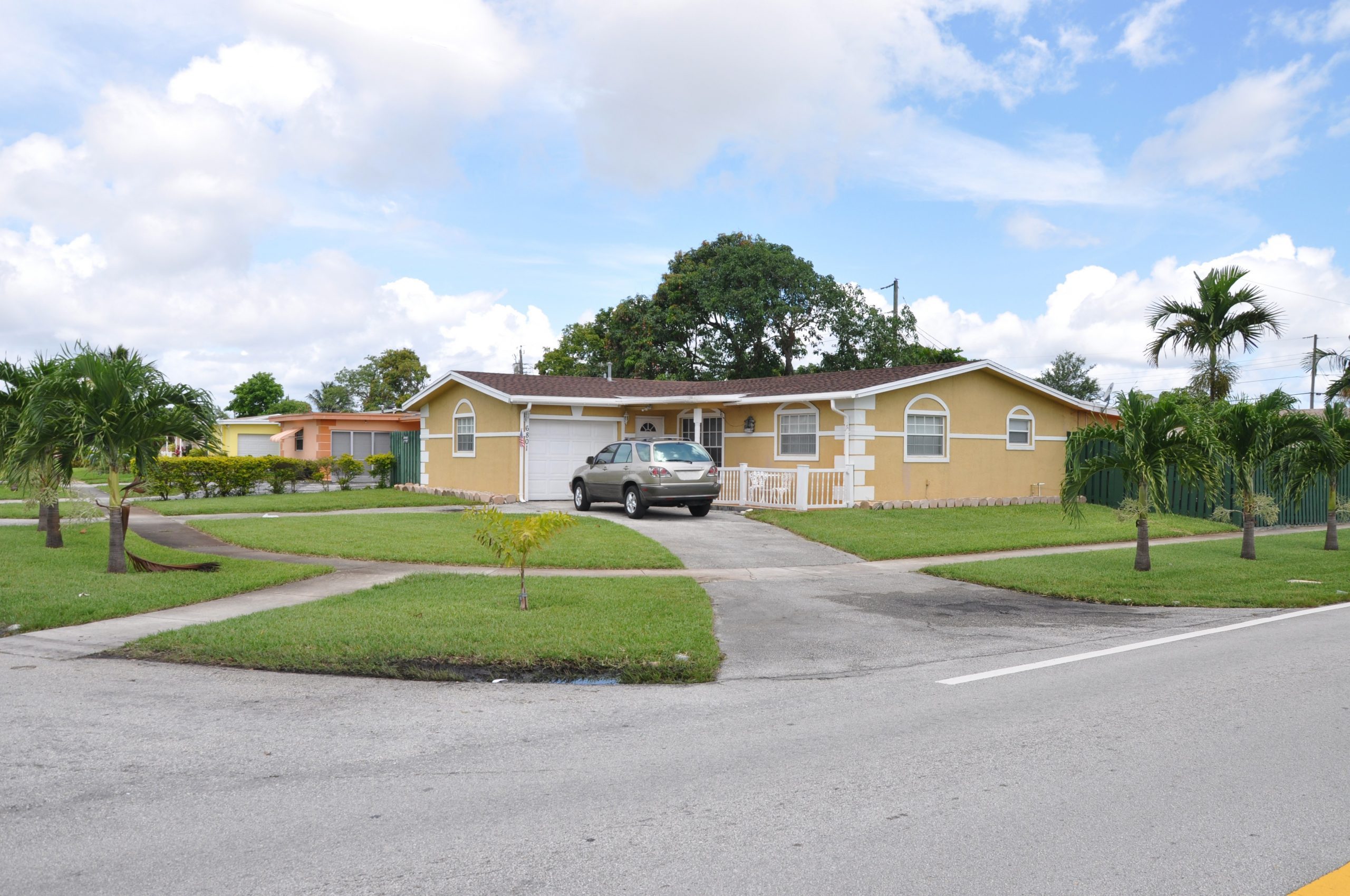 Suburban Neighborhood Corner Ranch Style House Palm Trees Car parked in driveway blue sky clouds day