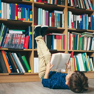 Boy laying on the floor with the feet up, reading a book against multi colored bookshelf in library. Education, Knowledge, Bookstore, Lecture.