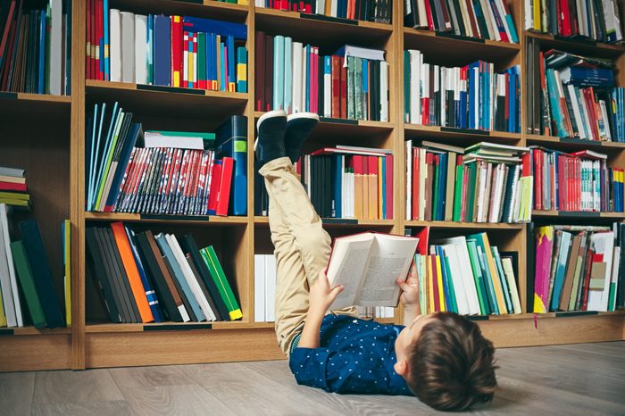 Boy laying on the floor with the feet up, reading a book against multi colored bookshelf in library. Education, Knowledge, Bookstore, Lecture. Pupil holds a book in his hands.