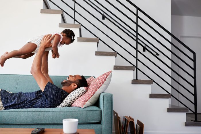 Father Lying On Sofa At Home Lifting Baby Daughter Into Air