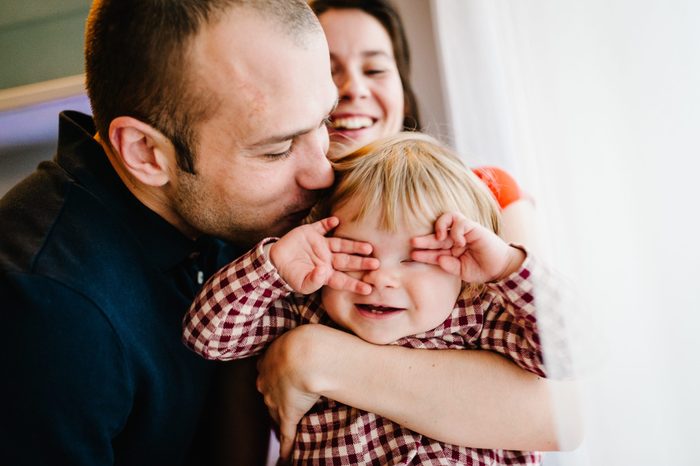 The father, mother holds little girl on background white wall indoor. Portrait of dad and daughter. The concept of family holiday. She closed eyes with hands and showed teeth. upper half. Close up.