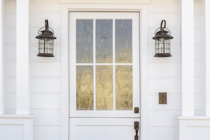 Cropped horizontal shot of a white door to a classic, white, family home in daytime. The door has intricate, etched, glass detail. Also see is light fixtures, columns, and a door bell. 