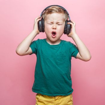 Stylish teen boy listening music in headphones and singing against pink background. School child listening loud music in wireless earphones and dancing.