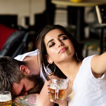 Take selfie to remember great event. Woman making fun of drunk friend. Man drunk fall asleep table and girl with full beer glass. Girl taking selfie photo drunk boyfriend. He appears too weak for her.