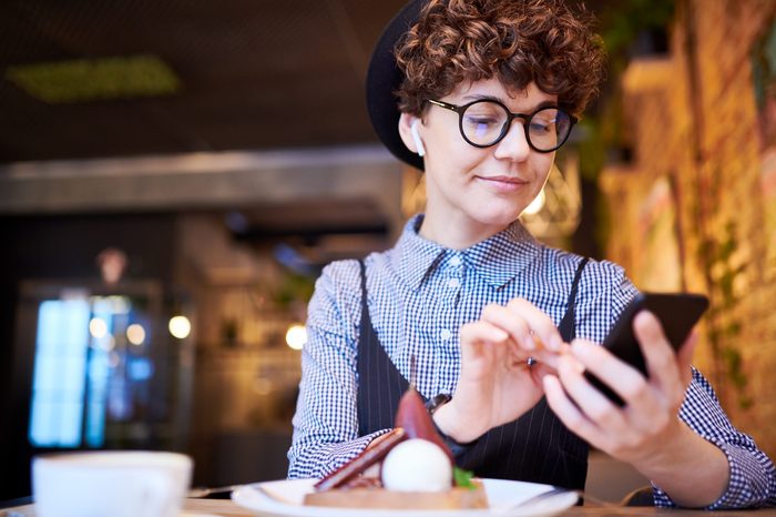 Young woman in eyeglasses and airpods scrolling in her smartphone through playlist while relaxing in cafe