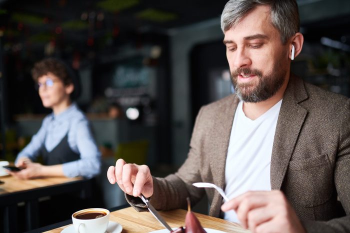 Contemporary young man with airpods and smart casual sitting in cafe and eating dessert for lunch