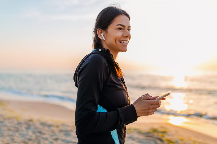 young attractive slim woman doing sport exercises on morning sunrise beach in sports wear, healthy lifestyle, listening to music on wireless earphones holding smartphone, smiling happy