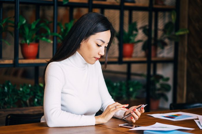 Positive kazakh businesswoman is thinking about tomorrow working plans and sending email to her boss using phone in the public place. Beautiful young female is doing her work in the cafe.