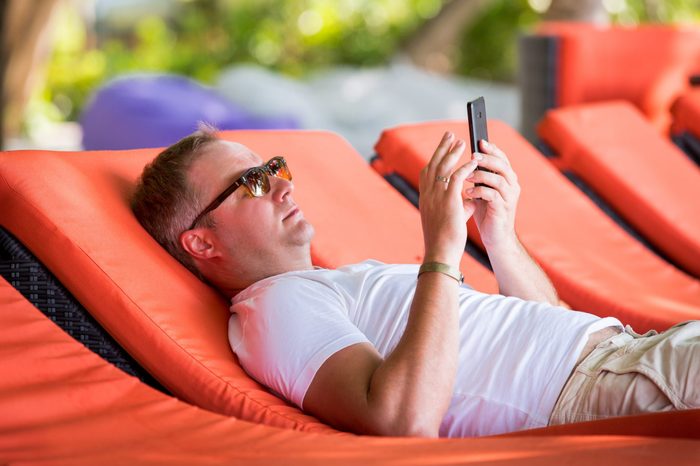 Man on a deck chair with smart phone on a summer day. Tourist calling by cell phone lying on the sunbed in a luxury hotel. Man relaxing in a tropical resort.