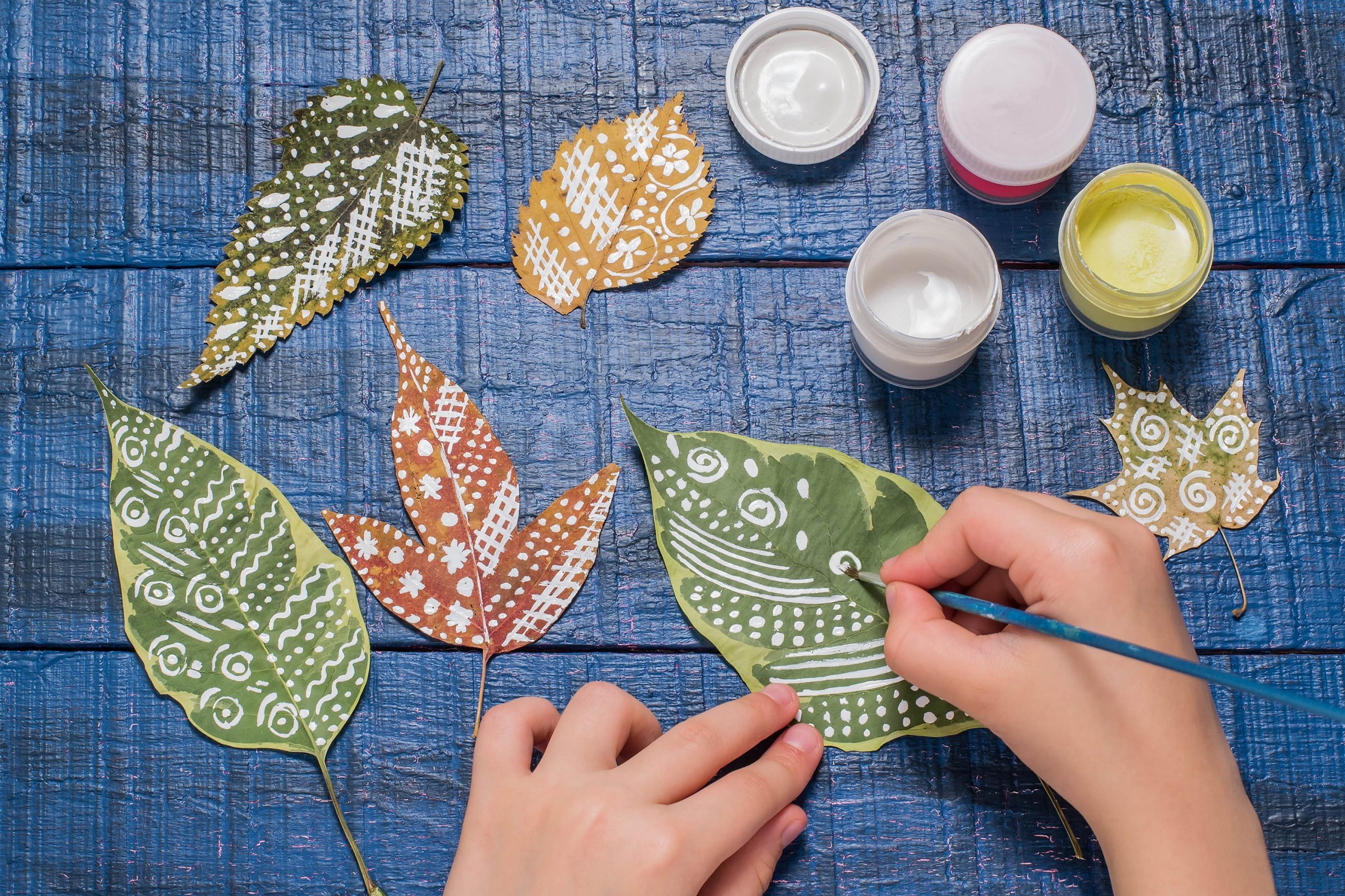 Girl paints patterns autumn leaf. Gouache, brush and various autumn leaves on a blue wooden table. Children's art project. Colorful Hand-painted on dry autumn leaves
