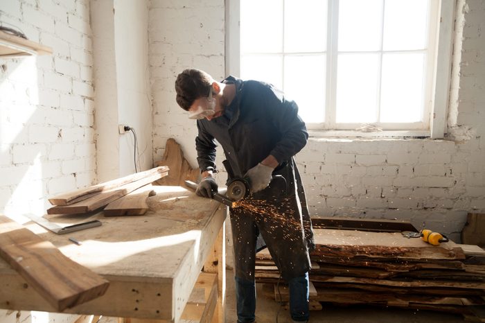 Worker cutting metal plank with angle grinder in workshop. Young man choosing skilled labor trades as first profession. Starting own small business, find side job in furniture manufacturing concept