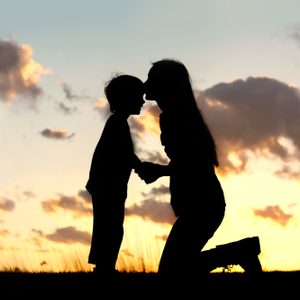 Silhouette of a young mother lovingly kissing her little child on the forehead, outside isolated in front of a sunset in the sky on a summer day.