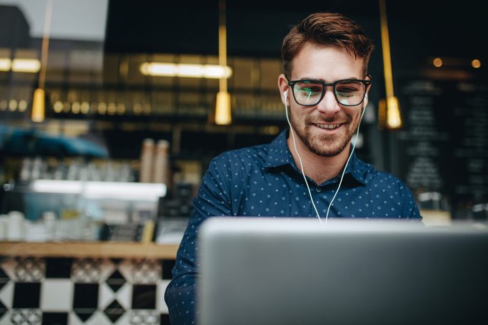 Businessman sitting in a restaurant listening to music while working on a laptop. Smiling man wearing earphones managing business work on laptop sitting in a cafe.