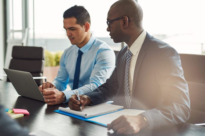 Two experienced business executives in a meeting seated at a table discussing paperwork and information on a laptop computer, one Hispanic, one African American