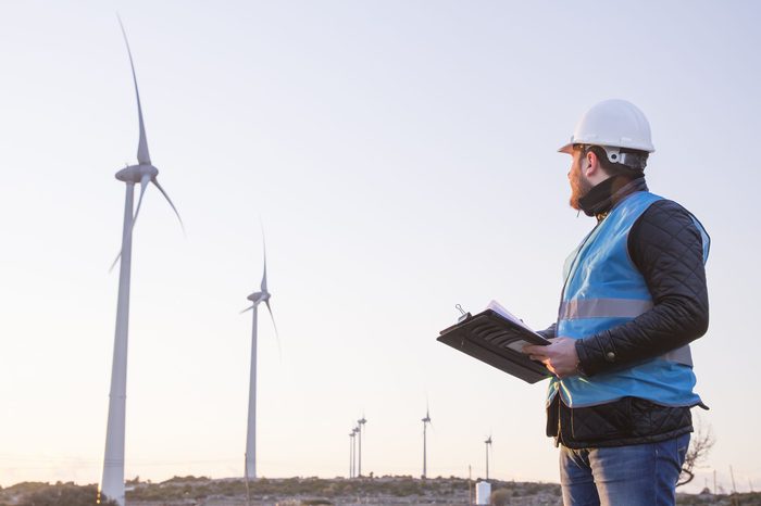 Young Handsome Beard Male Engineer in Wind Mill Power Generator Station with white safety hat and wind turbines on background