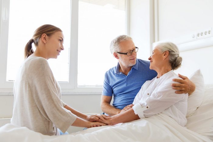 medicine, support, family health care and people concept - happy senior man and young woman visiting and cheering her grandmother lying in bed at hospital ward