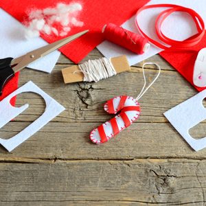 Christmas DIY craft of felt Christmas candy cane ornament, scissors, paper template, thread, needle, red and white felt pieces and scraps, filler, ribbon on old wooden background