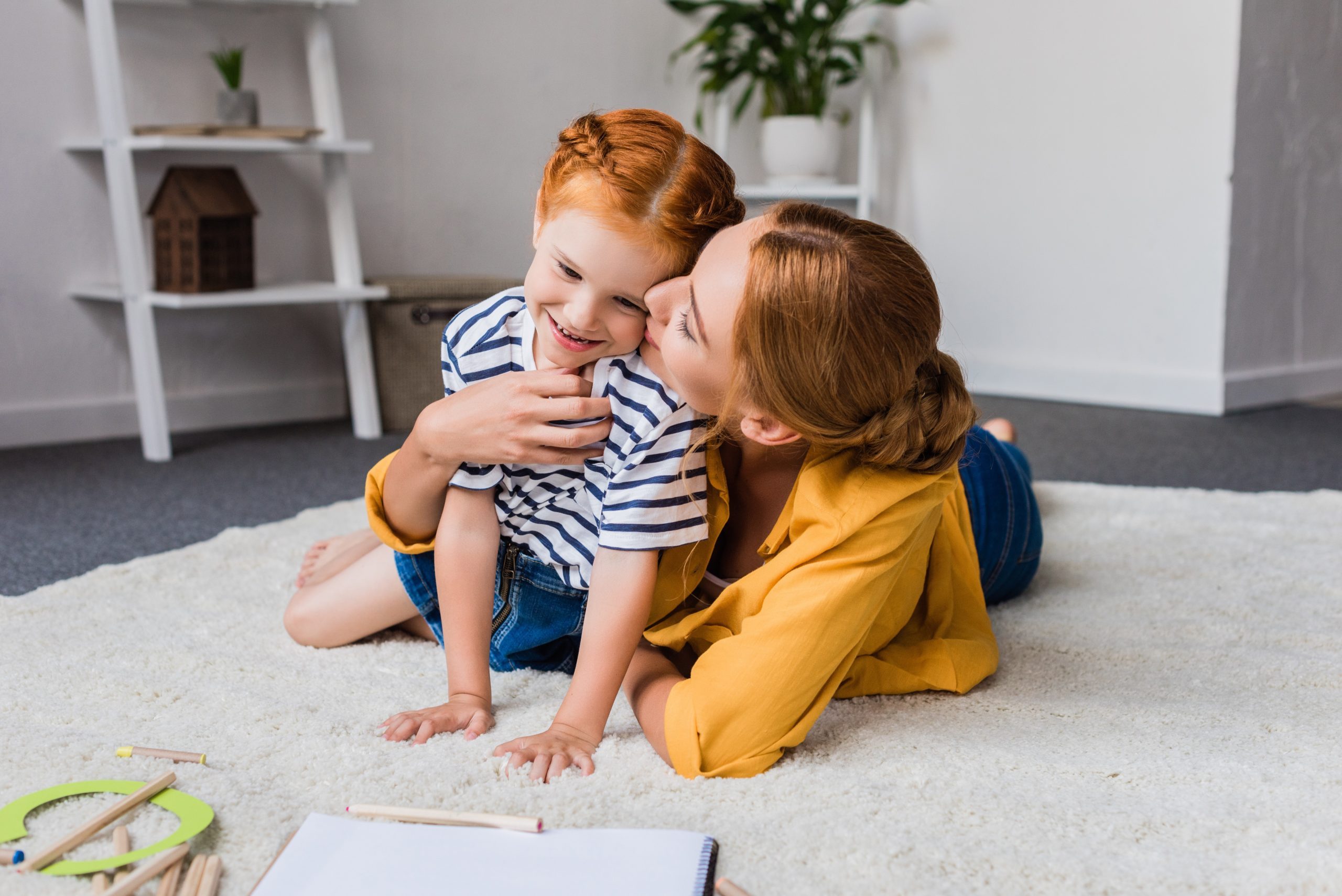 mother kissing her cute little daughter in cheek on floor at home