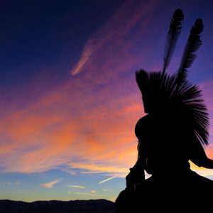 Native American Indian wearing head dress in silhouette against sunset background.