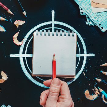 a hand holds a pencil over a small notebook that sits on top of chalk drawn crosshairs. scattered around is other pencils, shavings, and scraps of paper