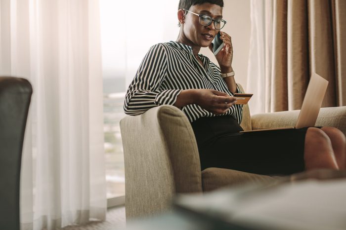 Female CEO sitting in hotel room with laptop and credit card talking over cell phone. African businesswoman doing online bill payments from hotel room.