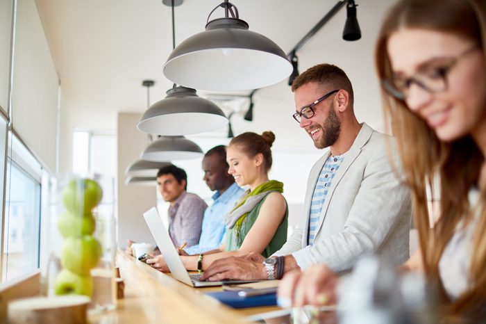 Row of casual business people smiling while working with laptops at standing workplace in modern light office