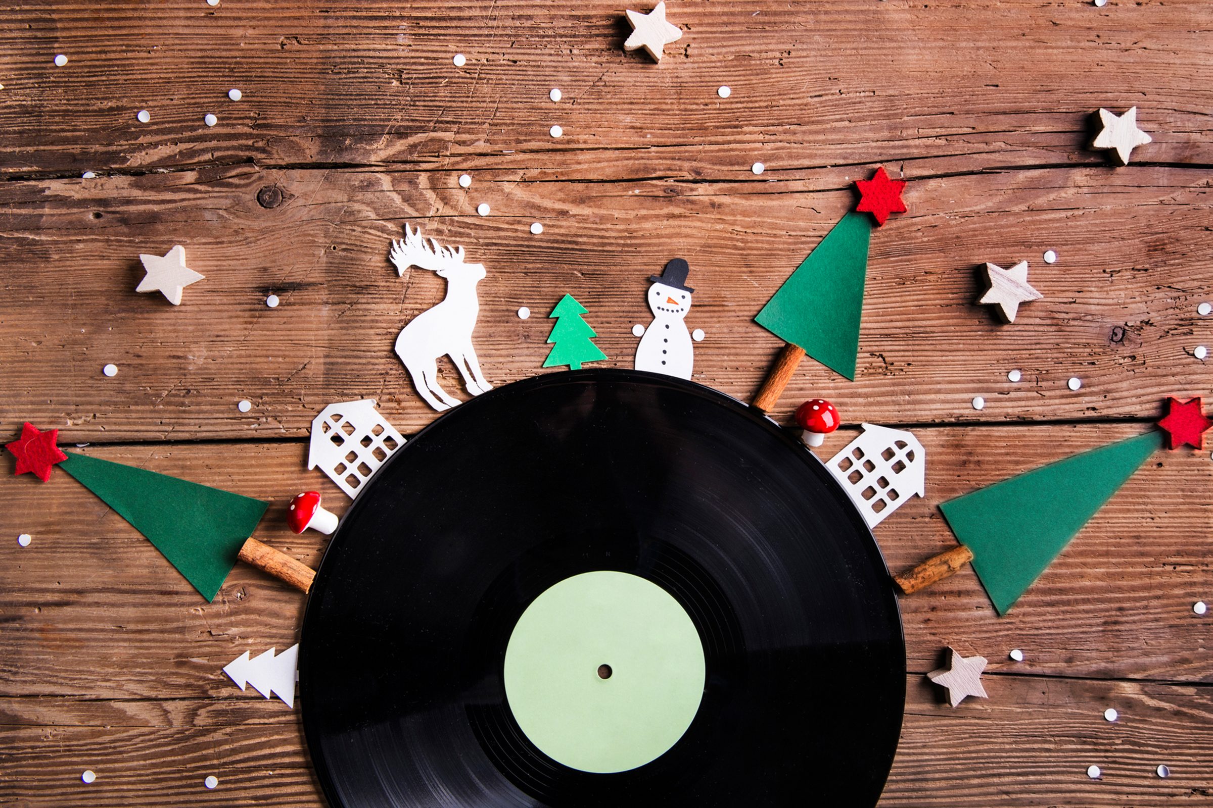 Flatlay of vinyl record with paper cutouts of Christmas trees and other Christmas items around the edge on a wood table background