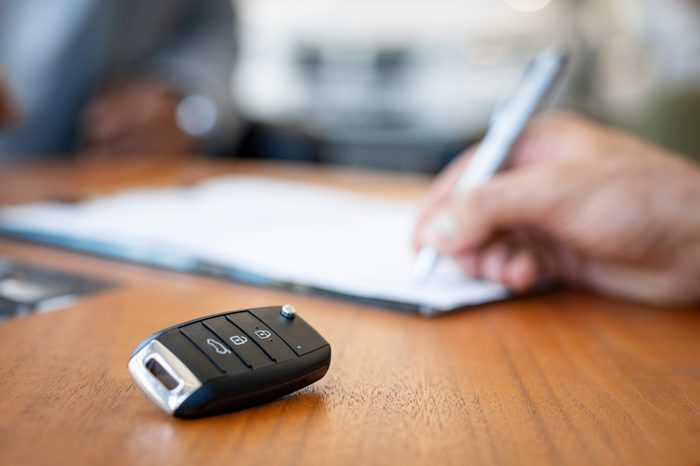 Car keys on desk with man signing purchase documents in background. Closeup of black modern car keys while hand complete the insurance policy or rental documents. Guy buying new car at dealership.