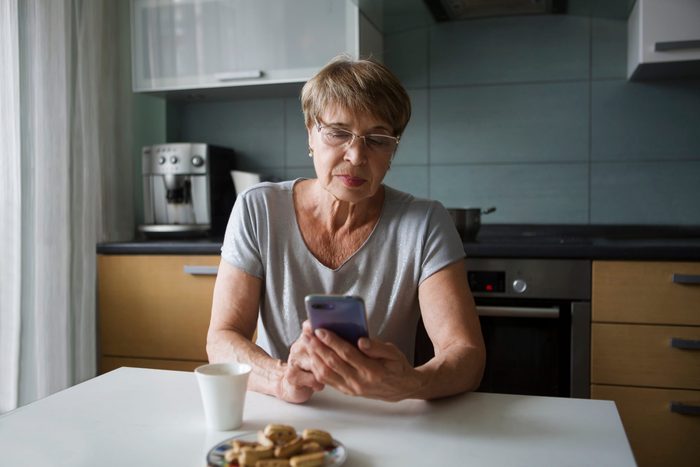 Portret happy senior woman looking at screen of mobile phone while in kitchen
